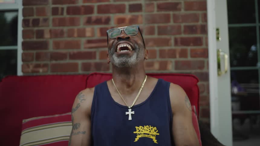 The video is a medium closeup of a bearded man with glasses and a dark tank top with a cross necklace sitting on a red cushioned chair outdoors he is seen smiling widely and then gesturing emphatically with his finger as he speaks