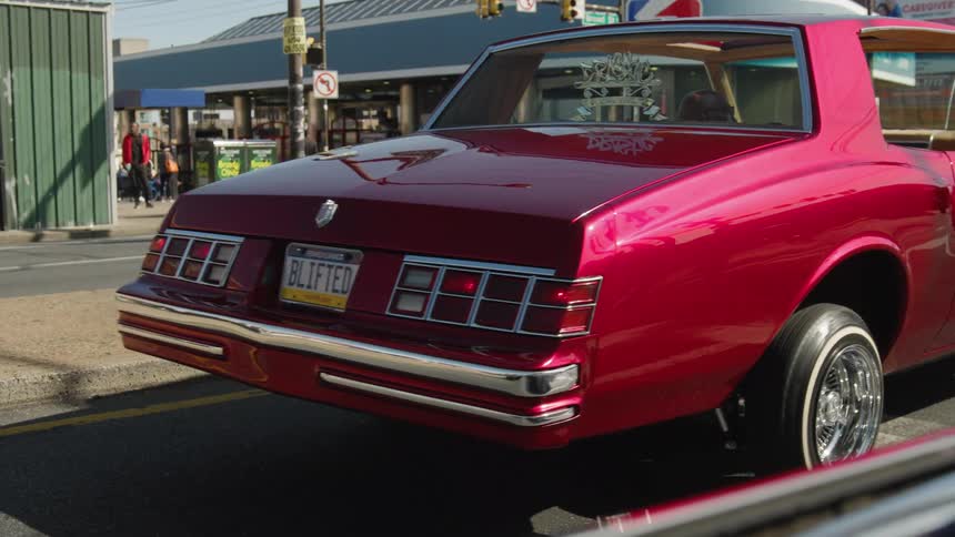 The video is a mediumcloseup shot of the rear end of a glossy cherryred classic car possibly a lowrider with shiny chrome accents the car has custom blifted license plates and is parked on a city street in daylight
