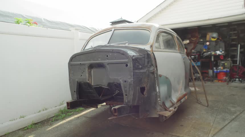 A low angle medium view of an old car undergoing restoration with the front fenders and grille removed sitting in a garage driveway on a bright overcast day