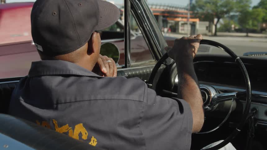 The video is an overtheshoulder shot of a man in a dark cap driving a classic convertible the man is wearing a dark work shirt with a yellow logo on the back
