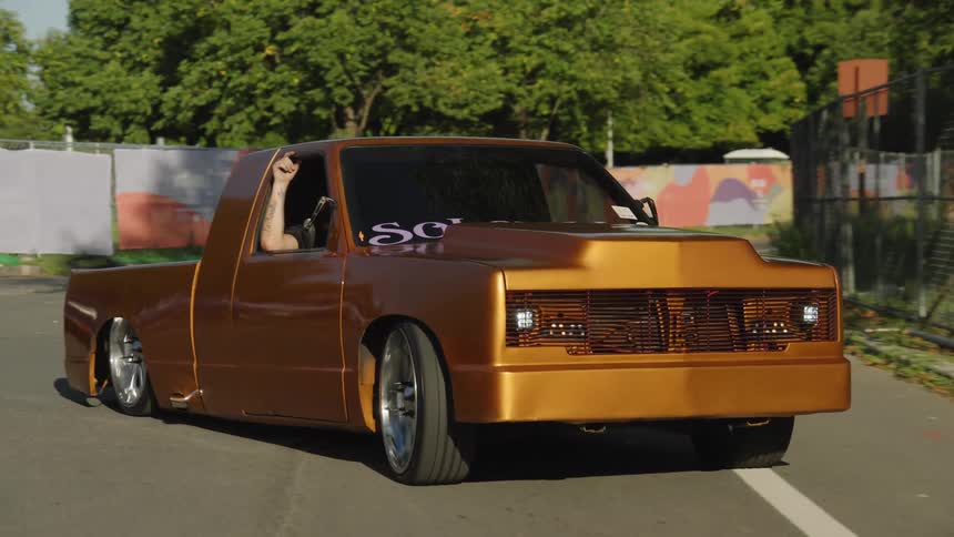 A highly modified lowslung pickup truck painted metallic copper drives along a road with trees and fences lining the sides approaching the camera and passing by other cars