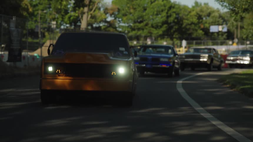The video shows a line of four or five classic vintagestyle american cars or muscle cars driving on a curved road through a park or wooded area the camera focuses on a large orangebrown vehicle in the foreground with its headlights on while