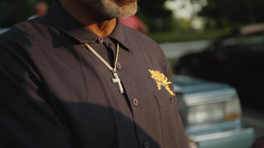 Person in dark embroidered shirt and gold necklace stands in front of classic car in outdoor area