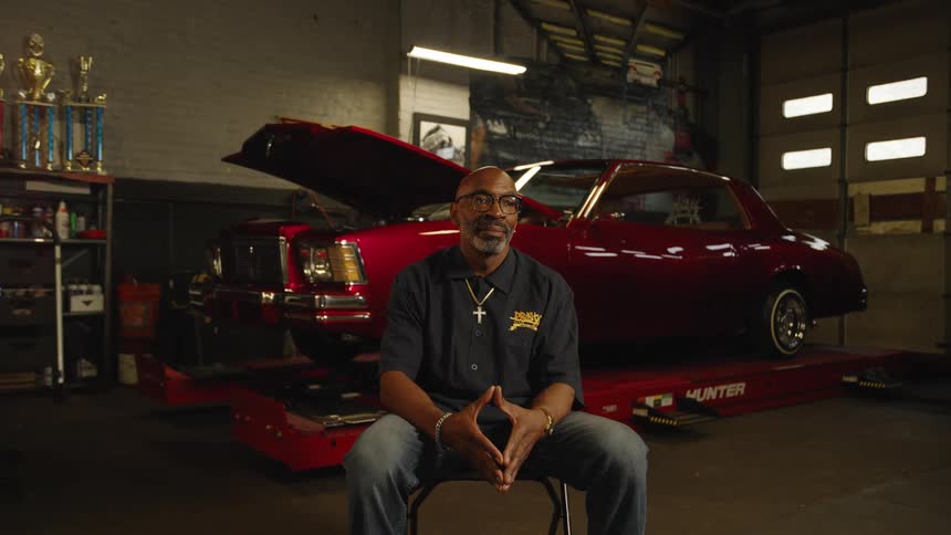 Man sits in an auto repair shop in front of a red vintage car on a lift