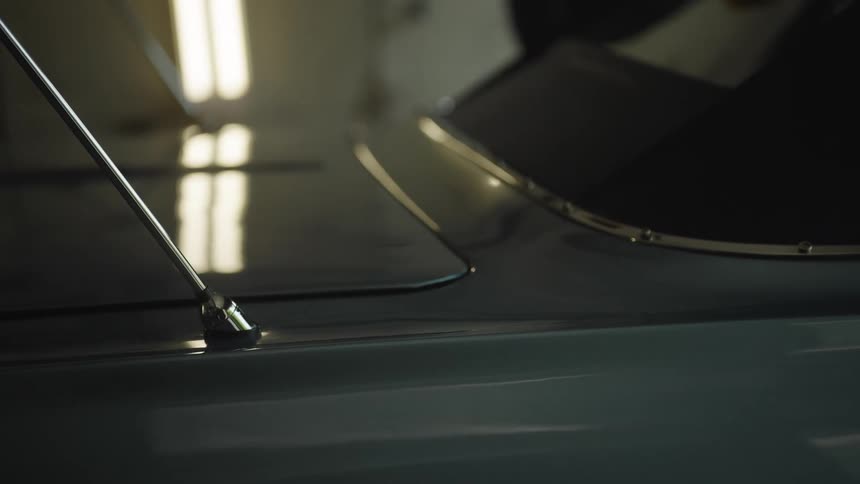 The video is an extreme closeup shot of a chrome antenna on the glossy dark blue or green surface of a classic car reflections of interior lights possibly fluorescent tubes are visible on the curved polished surface of the cars body