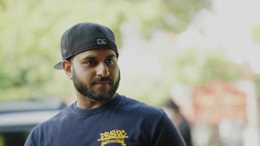 The video is a mediumcloseup of a young man with a beard wearing a dark blue tshirt and a black baseball cap standing outdoors he is looking offcamera and smiling slightly with green foliage and a blurry background suggesting a park or sub