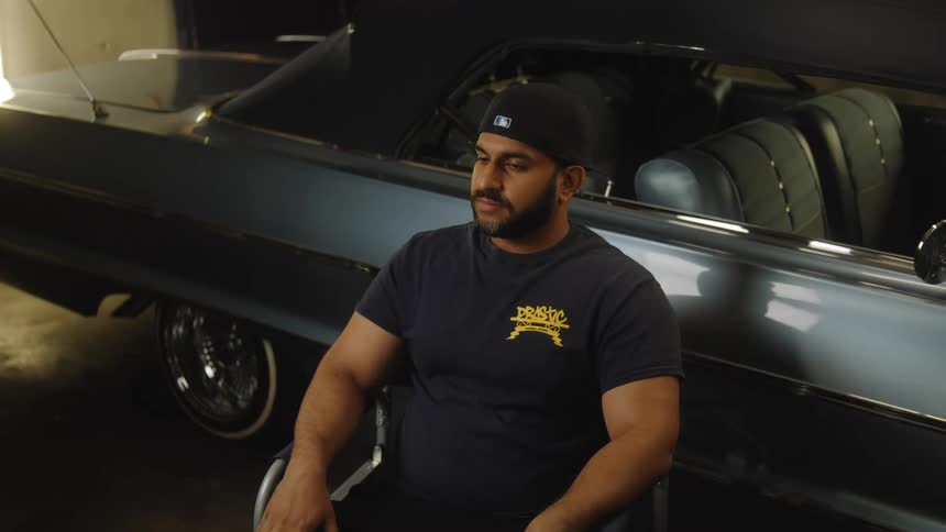 A man wearing a black tshirt and baseball cap sits in a chair facing the camera a dark convertible classic car is parked behind him the interior and exterior of the vehicle are visible and the scene takes place indoors in a garage