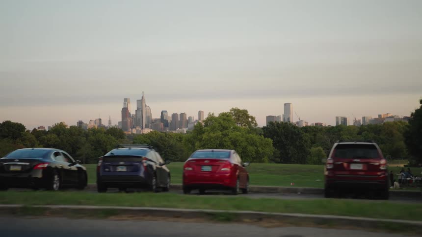 The video is a wide steady shot of a row of parked cars on the left side of a street facing a green treelined hill the skyline of a city likely philadelphia is visible in the distance above the trees