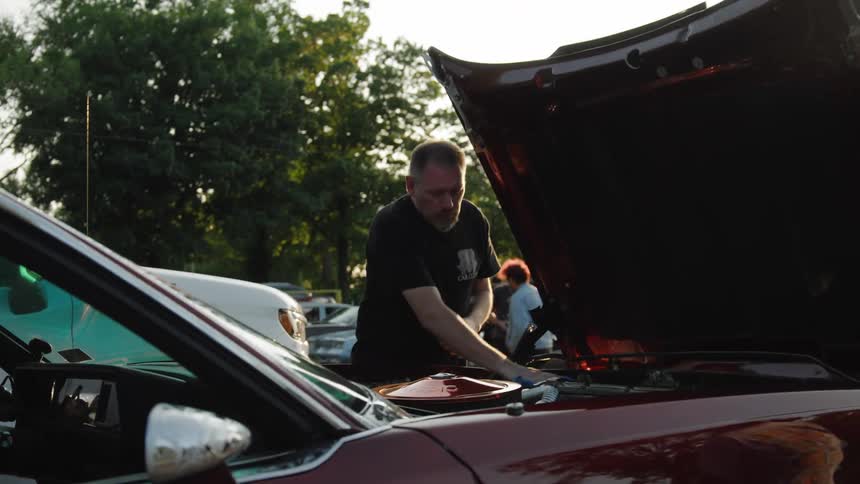 A man works on the engine of a vintage dark red car with the hood open at an outdoor event using a blue rag to wipe something under the hood