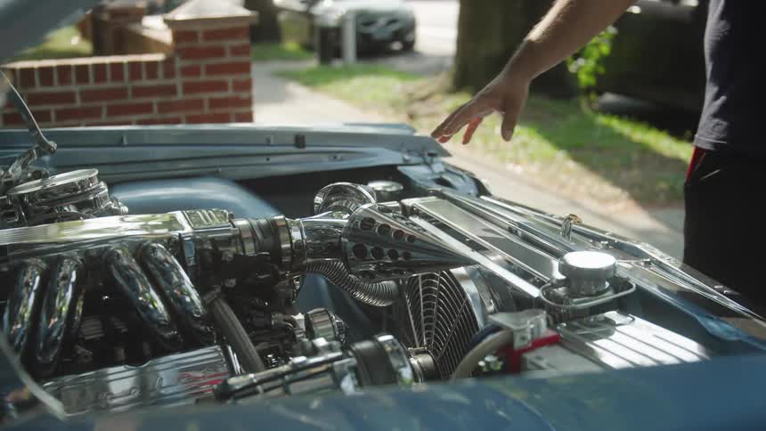 The video is a closeup of a highly polished chrome classic car engine with decorative pointed chrome intake funnels a mans hand enters the frame to gently touch the engines detailing as it is displayed outdoors in the sun