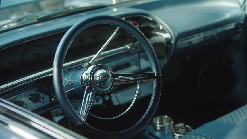 The interior of a classic car the large steering wheel with a chrome center and horn ring dominates the foreground the blue dashboard and instrument cluster are visible behind the wheel and the seat upholstery is textured