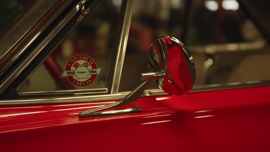 A close angled view shows the side mirror of a vintage red car with a circular sticker visible on the window next to the chrome trim and the mirror reflecting a warm indoor space