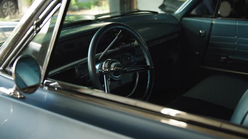 The interior of a classic car through the open side window the dark steering wheel with a chrome center is visible the dashboard and blue seats fill the cabin area and natural light enters the vehicle from the front and back