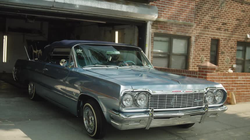 A classic blue convertible car slowly rolls forward out of a raised residential garage door positioned in front of a brick wall and house windows