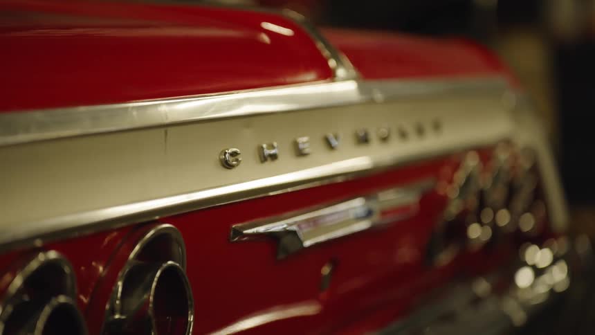 The rear of a vehicle shows red sheet metal and horizontal chrome strips the word chevrolet is visible on the center trim round tail lights are recessed into the body and the metal surfaces reflect surrounding light
