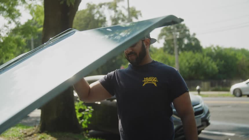 Man wearing a cap lifting the hood of a parked car in an urban area