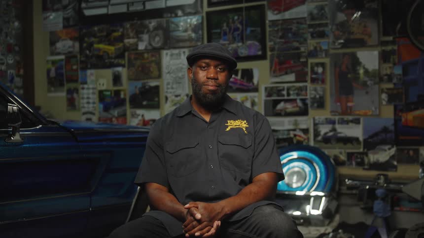 A medium view shows a man with a beard wearing a dark flat cap and a dark work shirt with a yellow logo sitting in a garage with a blue lowrider car to his left and a wall covered in photos behind him