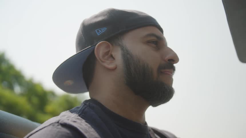 The video is a closeup profile shot of a mans face partially obscured by the brim of a dark baseball cap the man has a neatly trimmed beard and is looking intently offscreen to his right on a sunny day