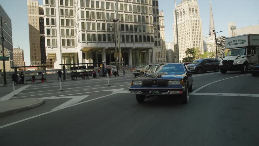 The video is an exterior shot of a brown lowrider car from the 1980s driving slowly past a bus stop and tall city buildings people wait at the bus stop and other modern cars are also seen driving on the city street