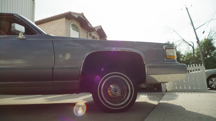 An eyelevel closeup shows the front fender and wheel of a classic grey sedan likely a cadillac with spoked wire wheels and whitewall tires the car is parked on a suburban street with a brick house and a white picket fence visible in the s