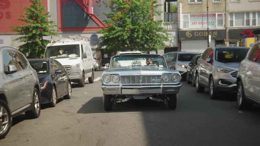 Man drives a classic car along a road lined with parked vehicles during the daytime