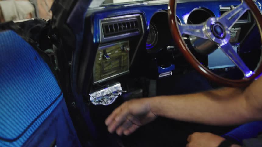 The video is an extreme closeup shot inside the dashboard of a classic car focusing on a persons hand as they adjust or activate a stereo unit the interior features vibrant blue patterned upholstery and a woodgrain detail on the dashboard