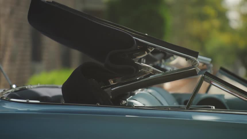 The video is a mediumcloseup of the mechanism and black fabric of a convertible roof moving up and forward the top is raising or lowering onto a glossy light bluegreen vintage car