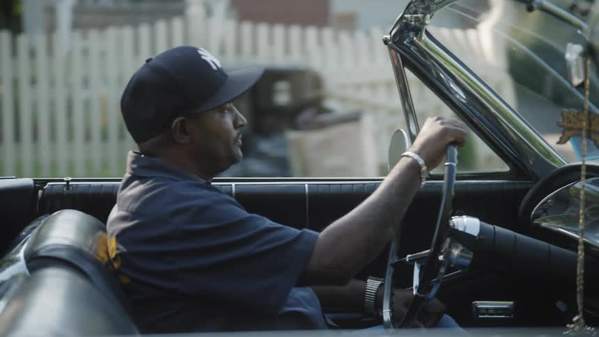 Man driving a black convertible in a residential area bordered by a wooden fence
