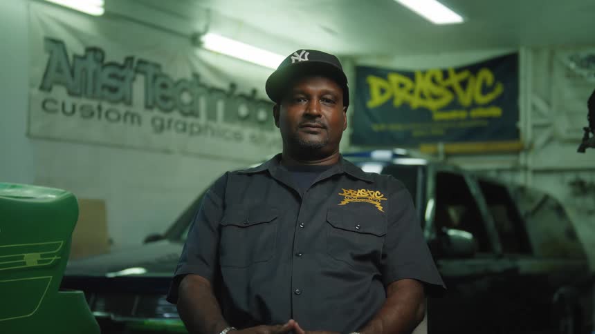 A medium view shows a man with a goatee wearing a dark work shirt with a yellow drastic logo and a yankees baseball cap as he looks directly at the camera in a dimly lit garage with cars and signs in the background