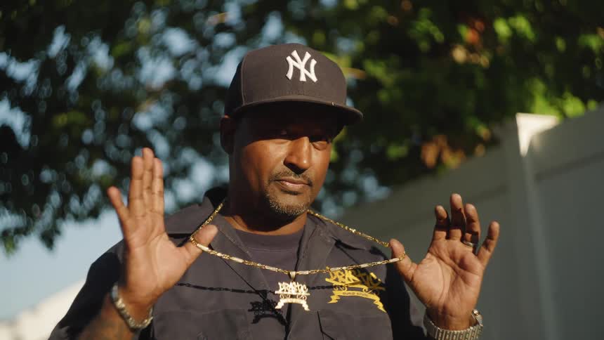 The video is a medium closeup of a man in a new york yankees baseball cap and a work shirt standing outdoors in bright sun he is holding up a large gold chain with a decorative pendant hanging around his neck looking directly at the camera