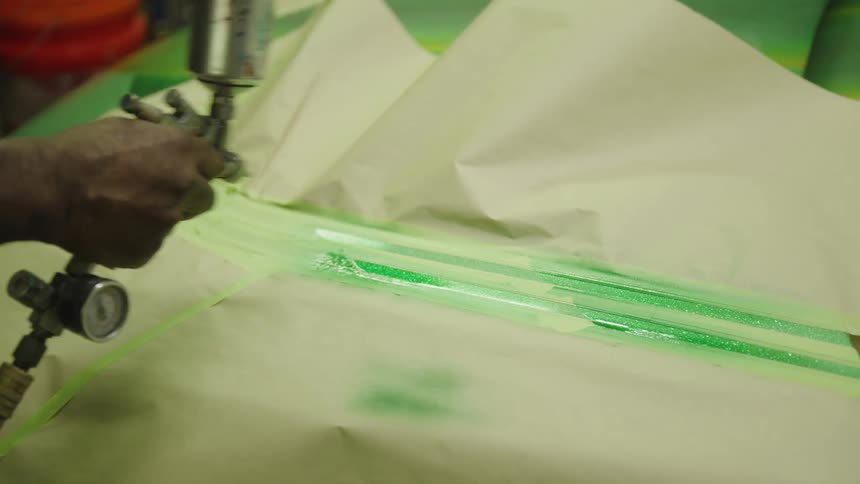 A persons hand holds a paint applicator the tool sprays a line of green color onto a white material surface a light brown paper masks the surrounding area and a gauge indicates the level of pressure in the system