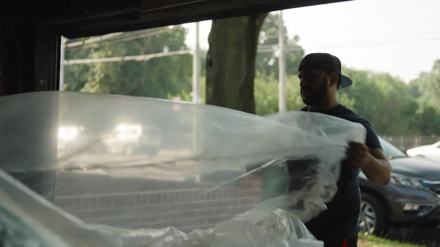 A man wearing a cap stands beside a car parked inside a garage opening and pulls back a large sheet of plastic covering the vehicle moving the sheet to the back of the car