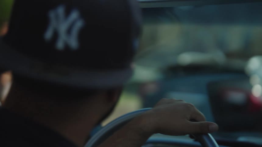 The back of a persons head wearing a baseball cap is visible as they firmly grip a steering wheel while driving a vehicle with the road and traffic visible out the windshield