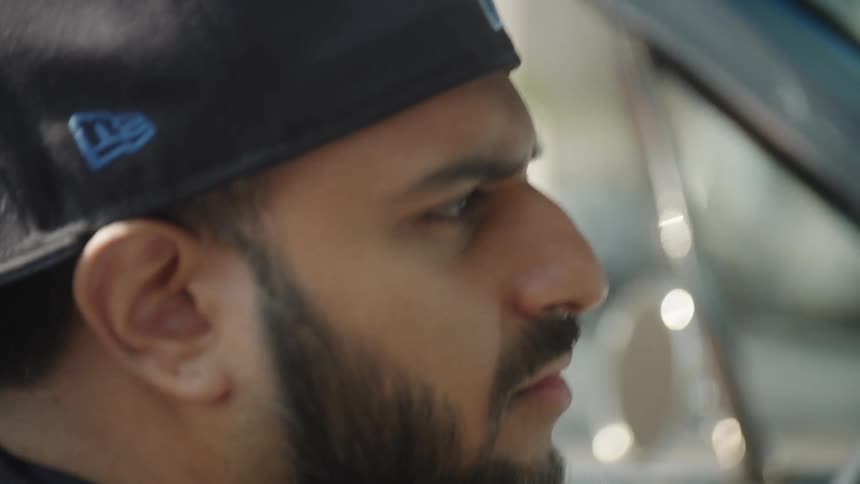 A closeup profile shot shows a man with a dark beard and a baseball cap worn backwards looking intently out of what appears to be a car window his expression is focused or serious and the background suggests he is either driving or a passenger