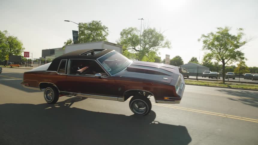 The video captures a classic darkcolored lowrider car driving slowly on a city street in bright sunlight with its front end lifted high off the ground by its hydraulic system the car features an open sunroof and is seen passing by a large stadi