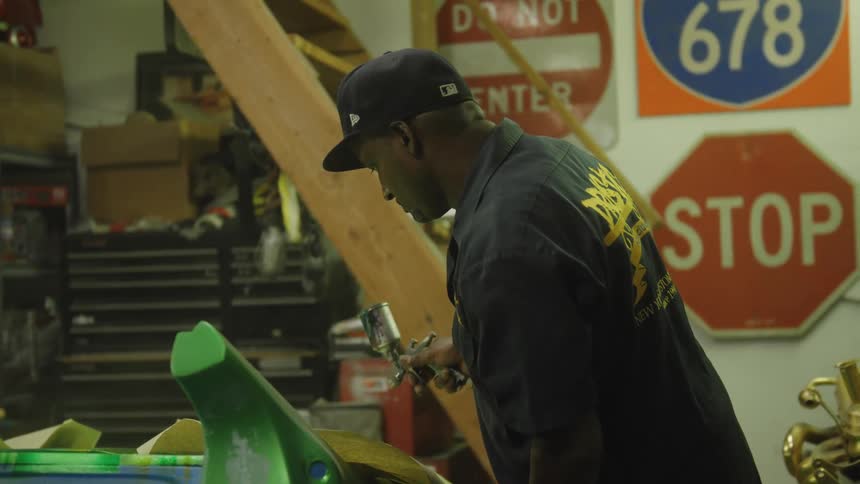 A man wearing a dark uniform and a baseball cap stands in an interior workshop holds a small spray apparatus applies a coating to a nearby curved surface and works amidst tools and traffic signs