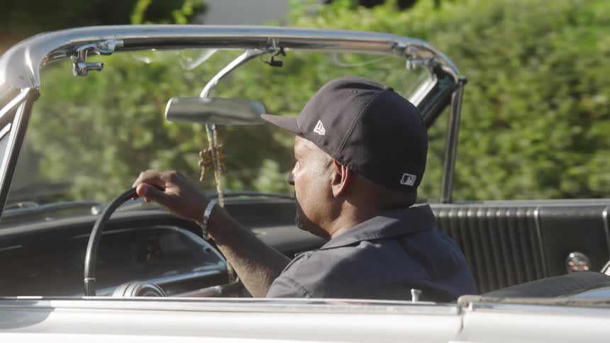 Man driving a white convertible in daylight wearing a dark cap