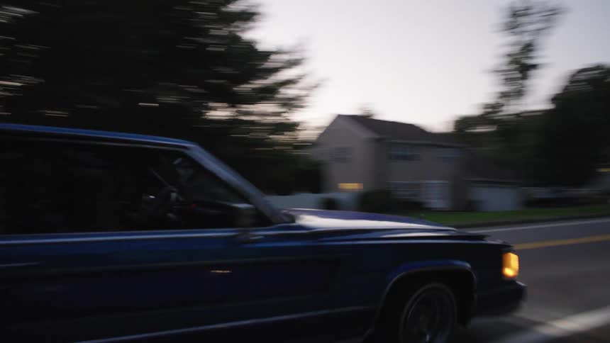 The video is a lowangle shot on a suburban or rural road at dusk or early night showing a darkcolored sedan driving past the camera the cars bright headlights and tail lights contrast with the dim light and trees and houses line the back