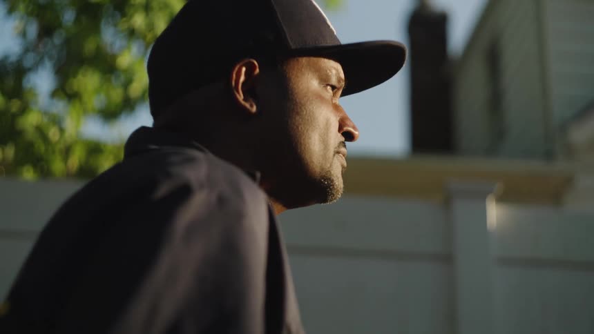A man in a dark baseball cap looks to the side in warm evening light with a pensive focused expression