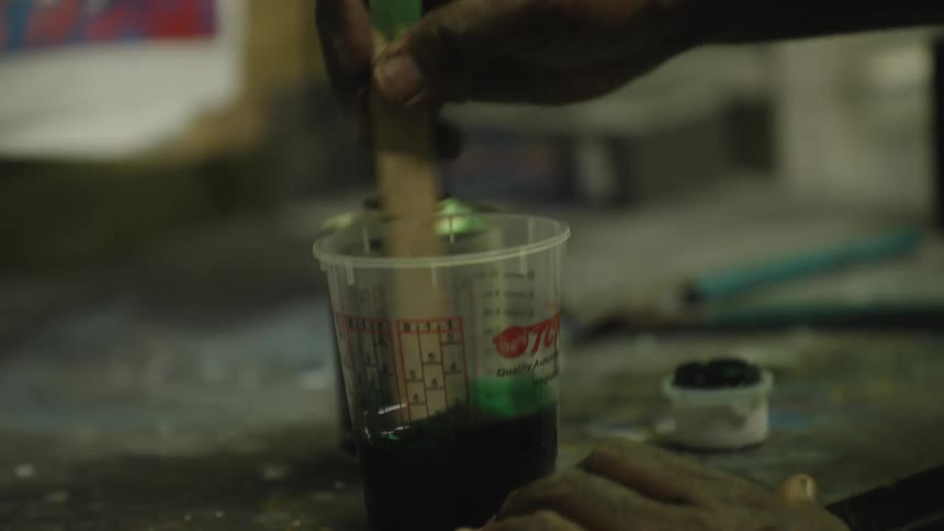 The video is a closeup shot of a hand mixing a dark liquid which appears to be paint or resin in a clear graduated plastic cup using a wooden stick as the stick is pulled out a drip of bright green liquid shows the mixing process is inco