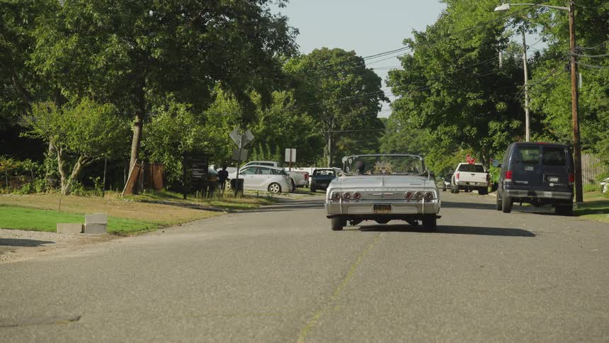A classic silver convertible lowrider with a chrome engine drives down an asphalt street lined with green trees and parked cars in a suburban neighborhood on a sunny day
