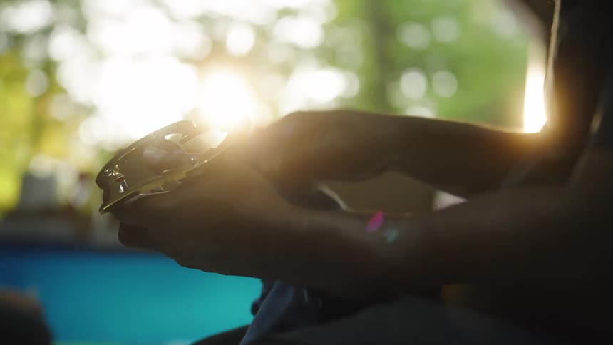 A persons hands hold a round chrome object and wipe it with a blue cloth the hands and object are silhouetted against an extremely bright background with strong sun glare and blurry green trees are visible in the distance