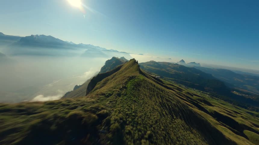 The video is a fastpaced fpv firstperson view drone shot soaring over a steep grassy mountain ridge with dramatic sheer cliffs below the ridgeline a valley is filled with a layer of mist or low clouds creating a spectacular mountain vi