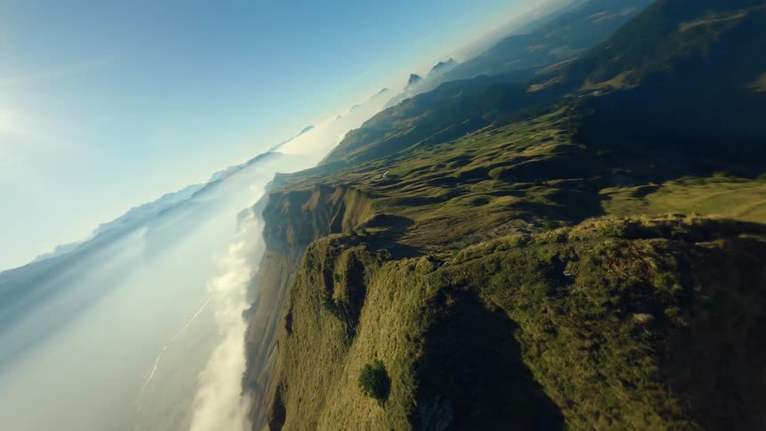 An aerial shot sweeps and dives over a steep grassy mountain ridge a person stands atop the narrow peak the background features multiple layers of mountains extending into the distance and the entire scene is illuminated by a bright sun