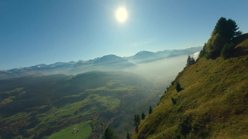 An extremely fast first person view drone shot flies over the edge of a steep grassy mountain ridge revealing a vast alpine valley filled with mist and fog with snowy peaks in the background under a bright blue sky with a strong sun