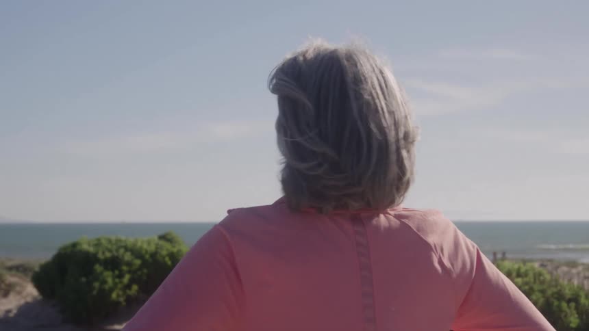 A grayhaired woman in a coral jacket stands at a seaside overlook with wind in her hair