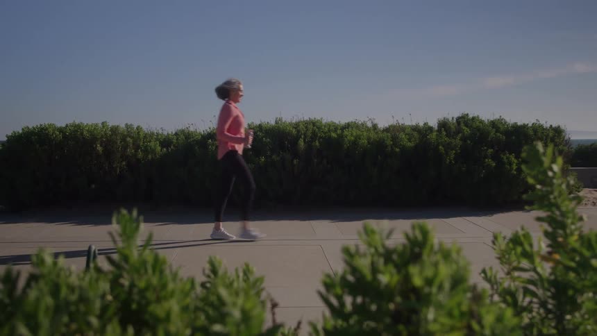 A woman with short gray hair wearing a pink shirt and black pants walks past a thick green hedge on a sunny sidewalk with green foliage in the foreground