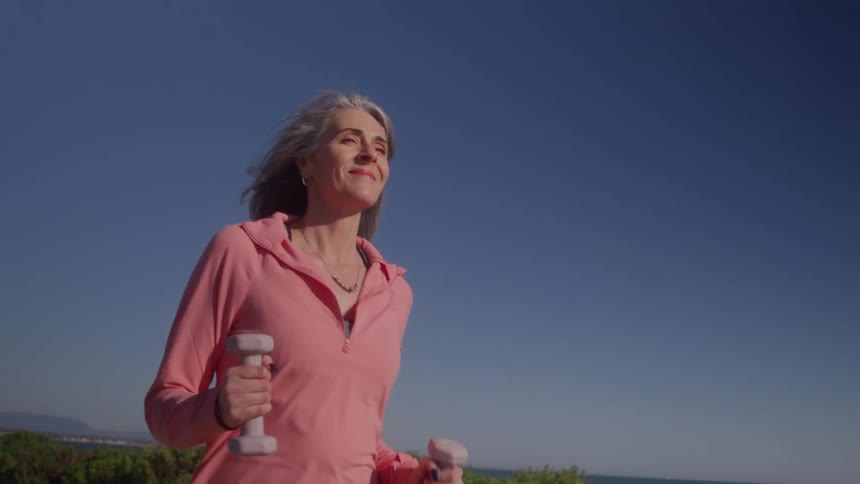 The video shows a smiling middleaged woman with graying hair wearing a pink hooded top as she jogs outdoors holding small hand weights in a sunny clearsky coastal environment the background features scrubby bushes and a glimpse of the oc