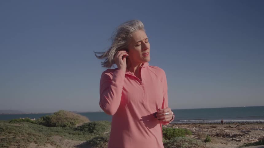 Grayhaired woman puts on earphones while walking by the seaside