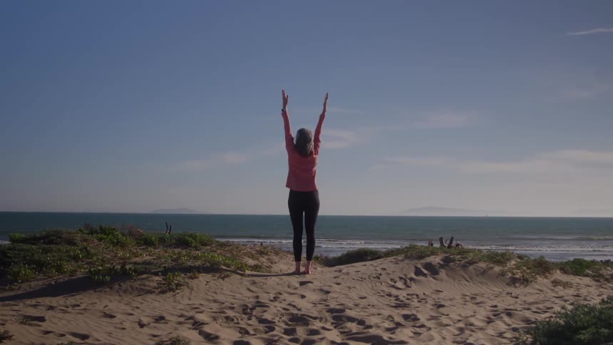 Woman stretching on sandy dunes by the ocean under a clear blue sky